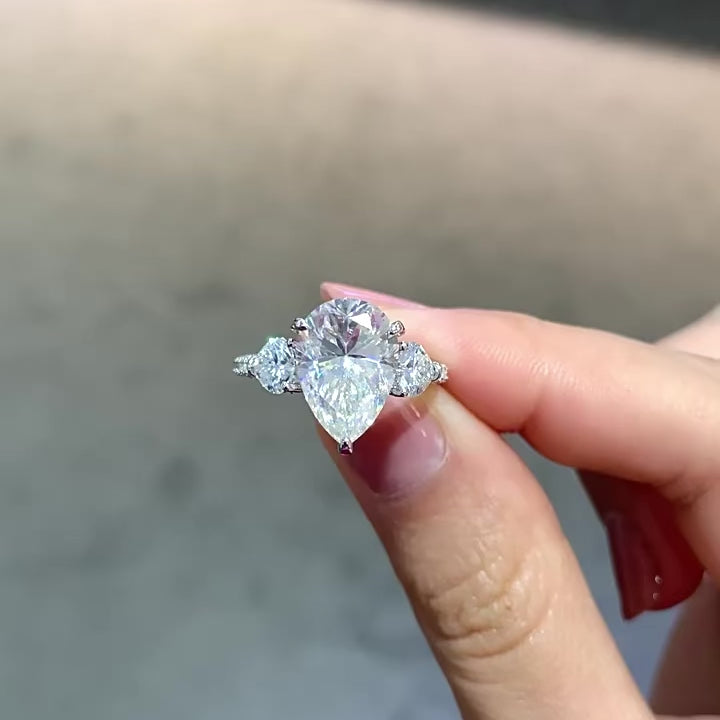 Sterling silver engagement ring with large pear-shaped moissanite center stone and heart-cut side stones, detailed with round moissanite on the band.